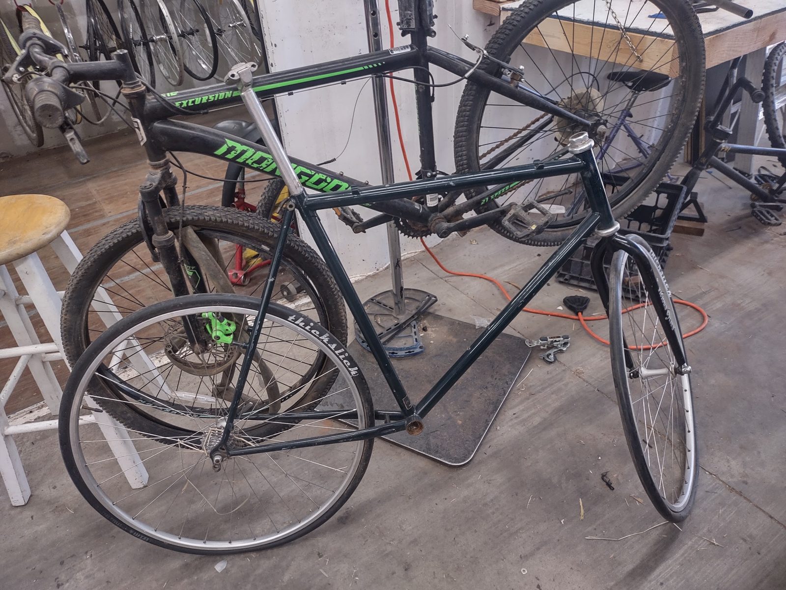 The green steel Novara frame with mismatched 700c wheels, leaning against the wall at the St. George Bicycle Collective