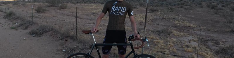 Isaac Lloyd standing behind the green singlespeed gravel bike on a dirt road in the desert, with red cliffs behind