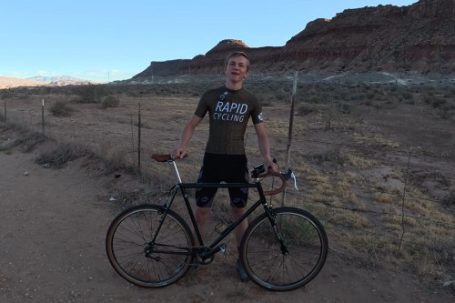 Isaac Lloyd standing behind the green singlespeed gravel bike on a dirt road in the desert, with red cliffs behind